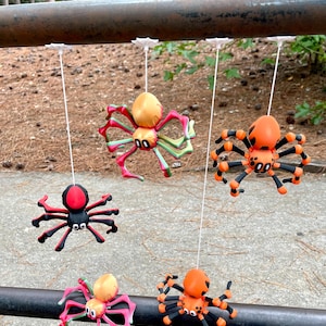 May include: Five colorful spider decorations hanging from a metal bar. The spiders are in various colors, including orange, black, red, and pink, with detailed legs and bodies. They are suspended by white strings, perfect for Halloween or spooky decor.