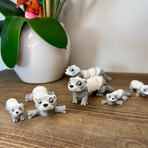May include: A collection of six gray and white ferret figurines with black eyes. The figurines are posed in different positions, including sitting, standing, and lying down. The figurines are on a wooden surface.