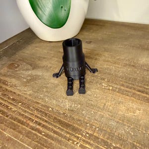 Peut inclure: Une cl&eacute; &agrave; douille noire de 10 mm avec des bras et des jambes attach&eacute;s, ce qui lui donne une apparence de dessin anim&eacute;. La cl&eacute; est pos&eacute;e sur une surface en bois, avec un pot blanc et une plante verte en arri&egrave;re-plan.