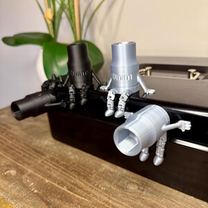 Peut inclure: Trois figurines de cl&eacute; &agrave; douille imprim&eacute;es en 3D, noires et argent&eacute;es. Chaque figurine a des bras et des jambes, et le texte "10mm" est visible sur deux d'entre elles. Les figurines sont sur une surface noire, avec une plante en arri&egrave;re-plan.