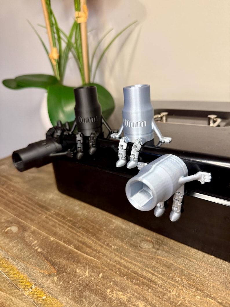 Peut inclure: Trois figurines de cl&eacute; &agrave; douille imprim&eacute;es en 3D, noires et argent&eacute;es. Chaque figurine a des bras et des jambes, et le texte "10mm" est visible sur deux d'entre elles. Les figurines sont sur une surface noire, avec une plante en arri&egrave;re-plan.