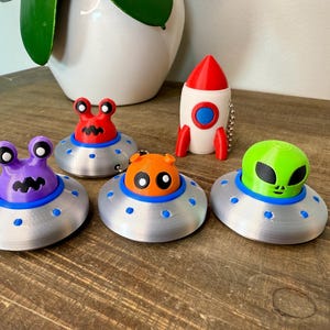 May include: Five colorful alien figurines on silver flying saucer keychains. The aliens are red, purple, orange, and green. A white rocket keychain is also included.