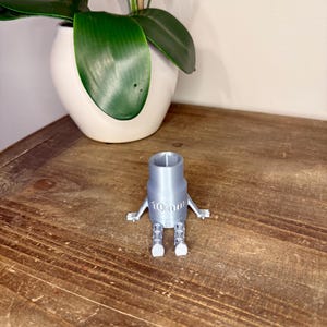Peut inclure: Une cl&eacute; &agrave; douille grise imprim&eacute;e en 3D, avec des bras et des jambes, portant l'inscription "10mm". L'outil est pos&eacute; sur une surface en bois, avec un pot en c&eacute;ramique blanc et une plante verte en arri&egrave;re-plan.