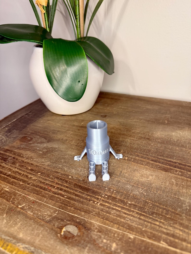 Peut inclure: Une cl&eacute; &agrave; douille grise imprim&eacute;e en 3D, avec des bras et des jambes, portant l'inscription "10mm". L'outil est pos&eacute; sur une surface en bois, avec un pot en c&eacute;ramique blanc et une plante verte en arri&egrave;re-plan.