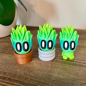 K&ouml;nnte beinhalten: Drei gr&uuml;n-blaue, gradientfarbene Figuren, die wie Sukkulenten aussehen, mit schwarzen Augen. Die Figur links befindet sich in einem braunen Topf, die mittlere Figur in einem grauen Topf und die Figur rechts steht auf einer Holzoberfl&auml;che.