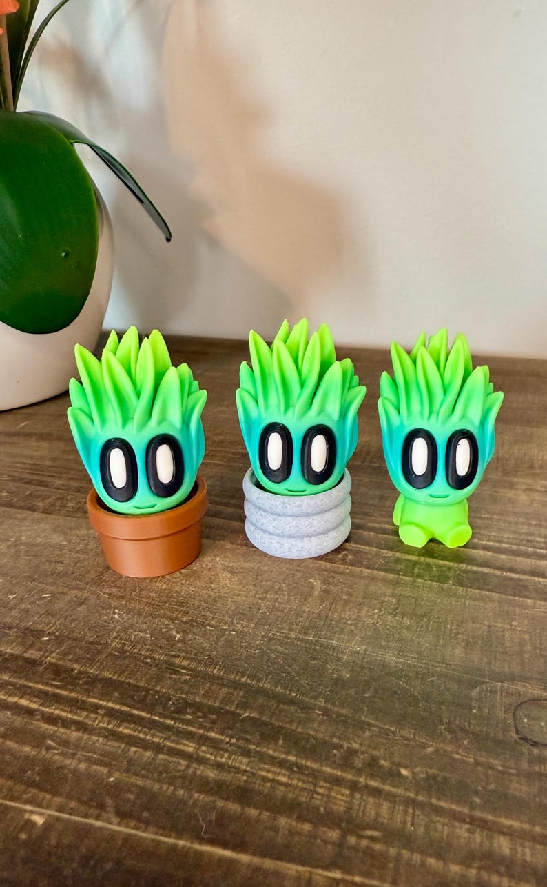 K&ouml;nnte beinhalten: Drei gr&uuml;n-blaue, gradientfarbene Figuren, die wie Sukkulenten aussehen, mit schwarzen Augen. Die Figur links befindet sich in einem braunen Topf, die mittlere Figur in einem grauen Topf und die Figur rechts steht auf einer Holzoberfl&auml;che.