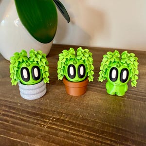 K&ouml;nnte beinhalten: Drei kleine gr&uuml;ne Pflanzenfiguren mit Gesichtern und verschiedenen T&ouml;pfen. Die Figuren sind aus glattem, gr&uuml;nem Material gefertigt und haben gro&szlig;e, wei&szlig;e Augen. Die T&ouml;pfe sind grau, braun und wei&szlig;.