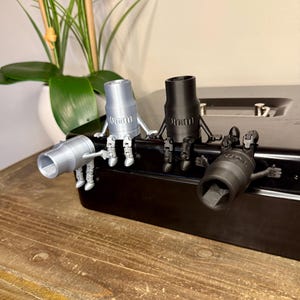 Peut inclure: Quatre figurines de cl&eacute;s &agrave; douille en argent, noir et gris, chacune avec des marquages de 10 mm. Les figurines sont pos&eacute;es sur une surface noire, avec une table en bois au premier plan et une plante en arri&egrave;re-plan.