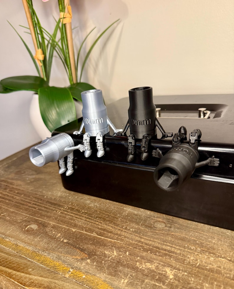 Peut inclure: Quatre figurines de cl&eacute;s &agrave; douille en argent, noir et gris, chacune avec des marquages de 10 mm. Les figurines sont pos&eacute;es sur une surface noire, avec une table en bois au premier plan et une plante en arri&egrave;re-plan.
