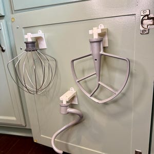 May include: Three kitchen mixer attachments, including a whisk, a flat beater, and a dough hook, are mounted on a light green cabinet door. Each attachment is held in place by a white holder, providing organised storage for baking tools.