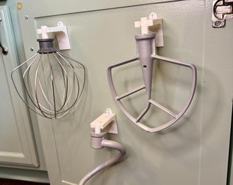 May include: Three kitchen mixer attachments, including a whisk, a flat beater, and a dough hook, are mounted on a light green cabinet door. Each attachment is held in place by a white holder, providing organized storage for baking tools.