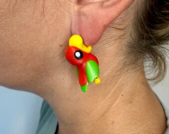 May include: A pair of colorful parrot earrings. Each earring features a parrot design with a red body, green wings, and a yellow crest. The parrot has a black and white eye and a yellow beak.
