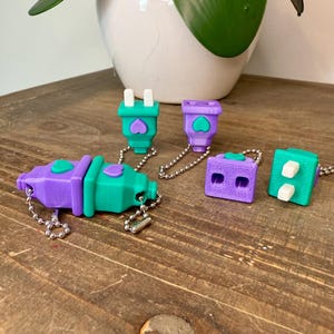 May include: A collection of colorful, 3D-printed plug-shaped keychains in purple and teal. Each keychain features a heart design and is attached to a small metal chain. The keychains are arranged on a wooden surface.