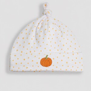 May include: A white baby hat featuring a pattern of small yellow stars and a knotted top. The front of the hat is embroidered with a single orange pumpkin with a green stem centred on the folded brim, providing a simple and clear design.