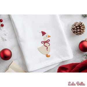 Könnte beinhalten: Weißes Handtuch mit einer gestickten Gans, die einen Weihnachtsmannhut und ein rotes Band trägt. Die Gans hat einen beigen Körper, orangefarbene Füße und einen Schnabel. Weihnachtsdekorationen, darunter rote Kugeln und ein Tannenzapfen, sind sichtbar.