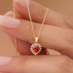 May include: A gold-colored necklace featuring a heart-shaped, red gemstone pendant. The pendant is surrounded by small, clear gemstones. The necklace is held by a person with light skin and long, manicured nails.