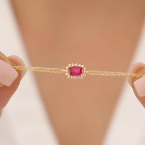 Könnte beinhalten: Ein zartes Armband aus einer goldenen Kette mit einem kleinen, quadratischen rosafarbenen Edelstein, der von einem Heiligenschein aus winzigen weißen Edelsteinen umgeben ist.