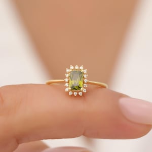 May include: A gold ring featuring a rectangular, olive-green gemstone surrounded by small, clear accent stones. The ring's band is thin and delicate, and the setting has a halo design. The ring is shown on a person's finger.