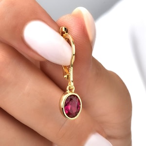 Könnte beinhalten: Goldene Creolen mit einem roten Edelstein, der herunterhängt. Der Edelstein ist rund und facettiert.