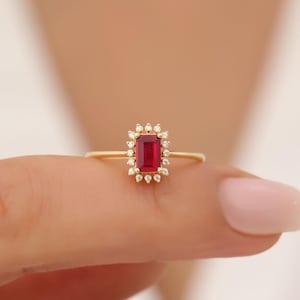 Könnte beinhalten: Ein goldener Ring mit einem roten rechteckigen Edelstein, der von kleineren Diamanten umgeben ist.