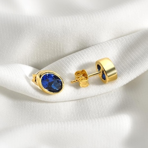 Könnte beinhalten: Ein Paar goldene ovale Ohrstecker mit blauen Edelsteinen.