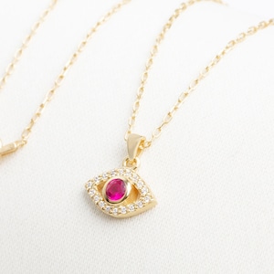 May include: A gold chain necklace with a pendant in the shape of an eye. The eye is surrounded by small clear stones and has a red stone in the center.