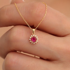 Könnte beinhalten: Eine goldfarbene Halskette mit einem Anhänger, der einen zentralen roten Edelstein und kleine, klare Steine ​​aufweist. Der Anhänger hat eine Blütenform. Die Halskette wird von einer Person gehalten.