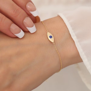 Könnte beinhalten: Ein goldenes Kettenarmband mit einem kleinen blauen bösen Blick-Anhänger. Der Anhänger ist oval geformt mit einem blauen Stein in der Mitte.