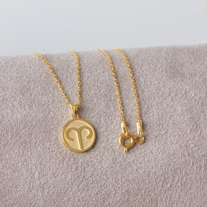 May include: A gold-colored necklace featuring a round pendant with the Aries zodiac symbol. The pendant hangs from a delicate chain with a lobster clasp. The necklace is displayed on a gray surface.