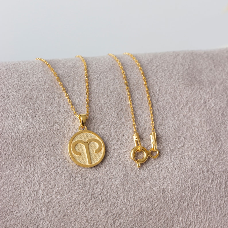 May include: A gold-colored necklace featuring a round pendant with the Aries zodiac symbol. The pendant hangs from a delicate chain with a lobster clasp. The necklace is displayed on a gray surface.