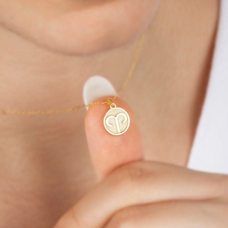 May include: A gold-colored necklace with a small, round pendant. The pendant features the Aries zodiac symbol in a minimalist design. The necklace is delicate and appears to be worn close to the neck, with the chain and pendant in sharp focus.