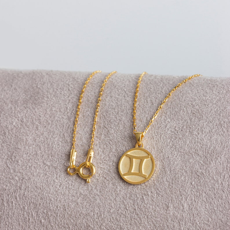 May include: A gold necklace with a round pendant featuring the Gemini zodiac symbol. The necklace is on a light brown fabric surface.
