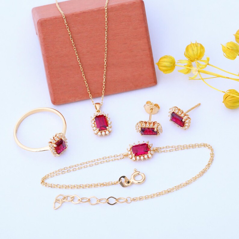 May include: A gold jewelry set with a red gemstone pendant, earrings, ring, and bracelet. The jewelry features a square-shaped red gemstone surrounded by smaller white gemstones.