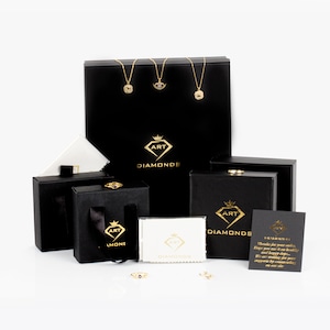 Op de afbeelding: Een verzameling zwarte juwelendozen met gouden letters die "ART DIAMONDS" lezen. De dozen zijn gerangschikt op een wit oppervlak. Er zijn ook twee kleine witte poetsdoekjes met dezelfde gouden letters. Een van de dozen heeft een bedankbriefje met de tekst "Bedankt voor uw bestelling. We hopen dat u het net zo leuk vindt als wij. We wachten op uw mooie recensies. We zijn blij met uw steun door onze sieraden te kiezen."