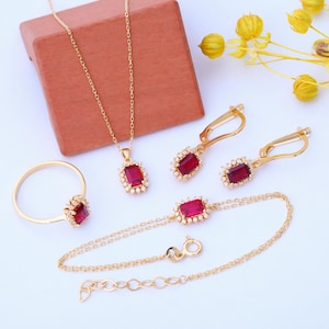May include: A gold jewelry set with a necklace, bracelet, ring, and earrings. Each piece features a red gemstone surrounded by smaller white gemstones.