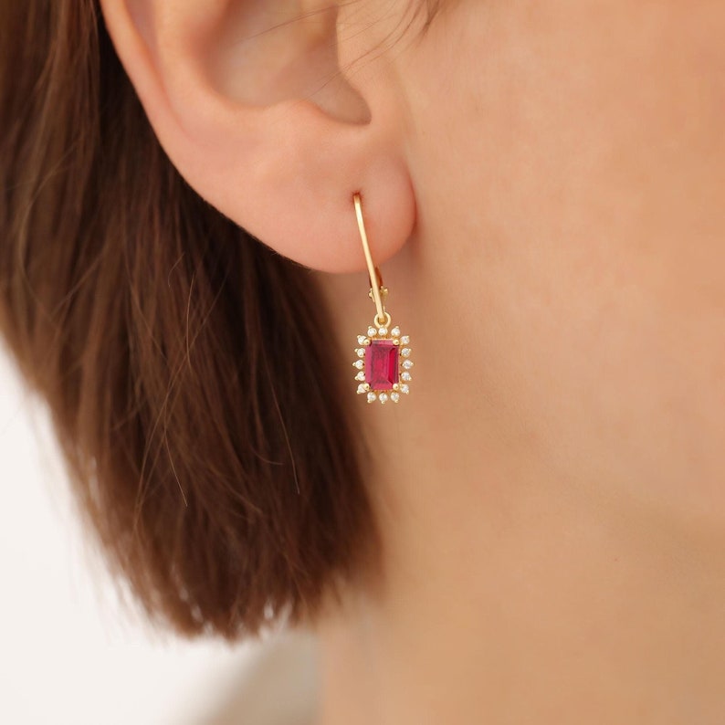 May include: Gold dangle earrings with a red gemstone surrounded by smaller clear gemstones.