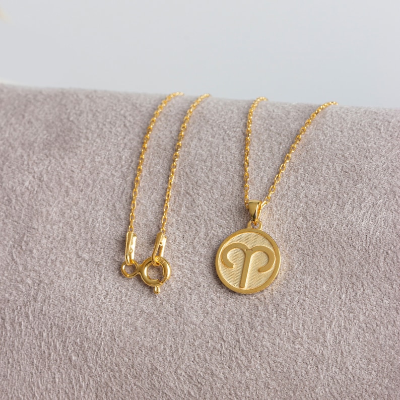 May include: A gold-colored necklace with a pendant featuring the Aries zodiac symbol. The pendant is circular with a textured background. The chain and clasp are also gold-colored. The necklace is displayed on a gray surface.