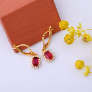 May include: A pair of gold earrings with a red gemstone and a halo of small clear stones.