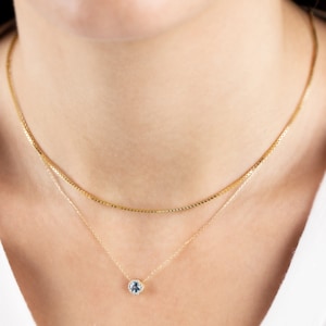 Op de afbeelding: Een gouden ketting met een delicaat hangertje met een ronde blauwe edelsteen omringd door kleinere heldere edelstenen.