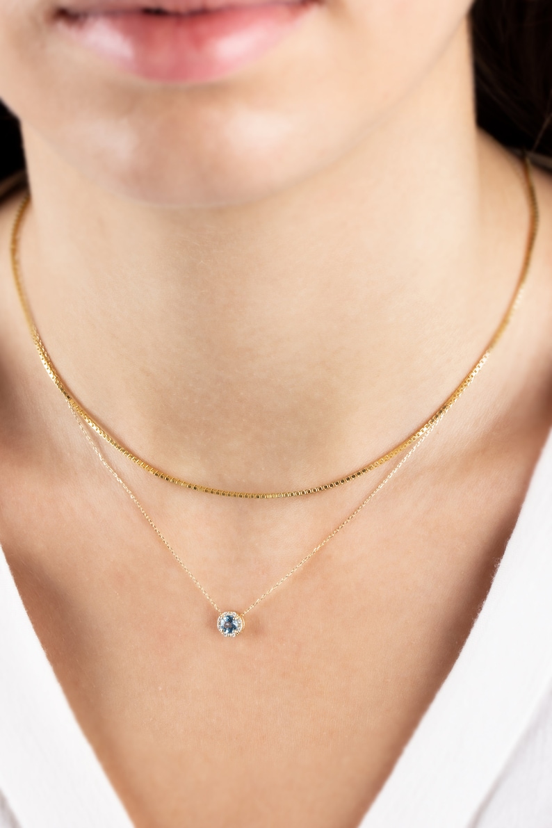 Op de afbeelding: Een gouden ketting met een delicaat hangertje met een ronde blauwe edelsteen omringd door kleinere heldere edelstenen.