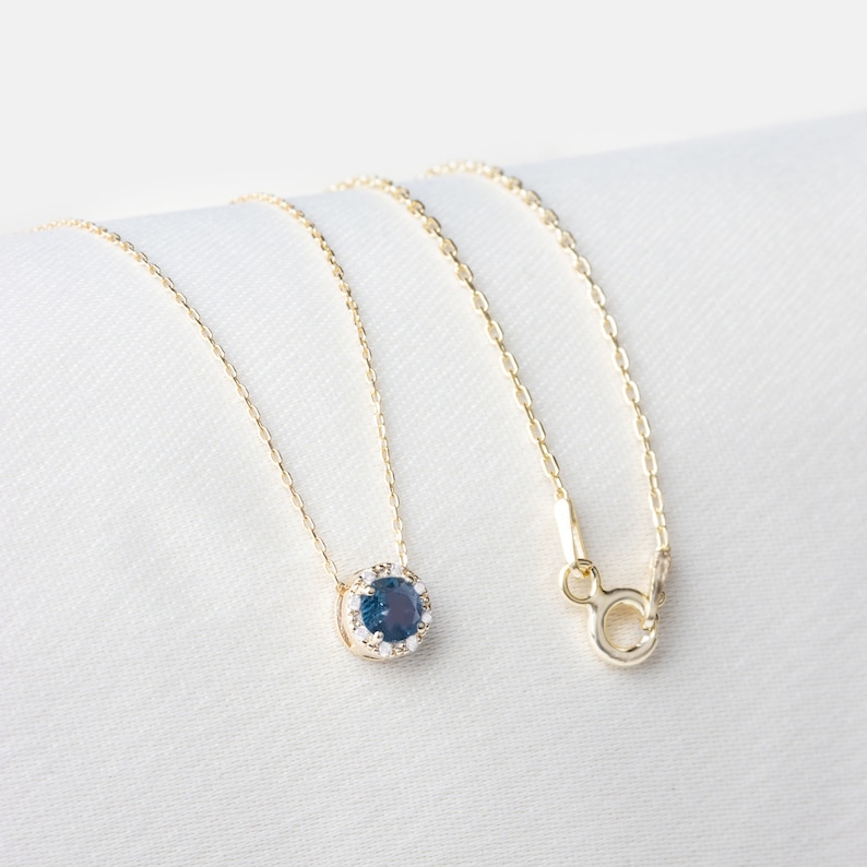 Op de afbeelding: Een gouden ketting met een blauwe edelsteen hanger omringd door kleinere heldere edelstenen.