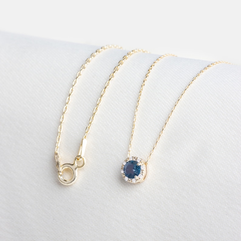 Op de afbeelding: Een gouden ketting met een blauwe edelsteen hanger omringd door kleinere witte edelstenen.