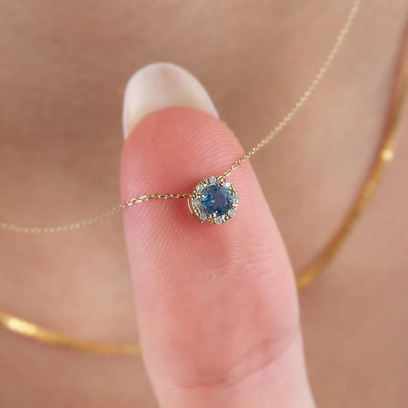Op de afbeelding: Een delicate gouden ketting met een blauwe edelsteen hanger omringd door een halo van kleinere diamanten.