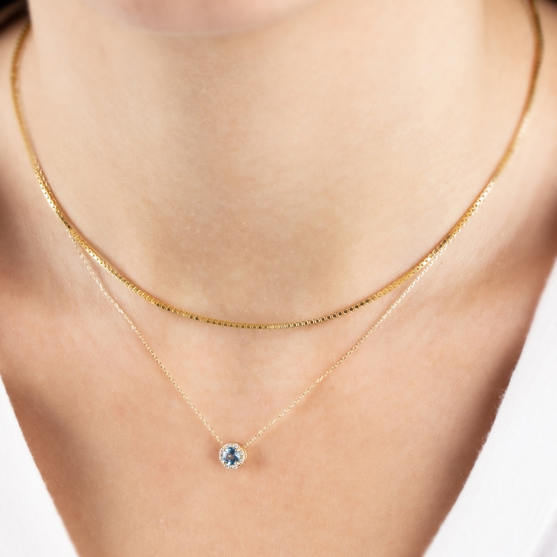 Op de afbeelding: Een gouden ketting met een klein, rond hangertje met een lichtblauwe edelsteen omringd door kleinere, heldere edelstenen.
