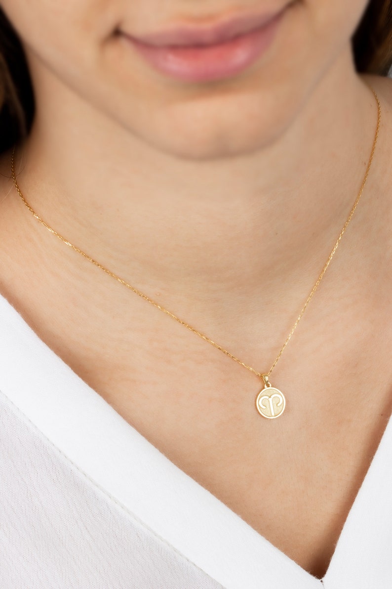 May include: A gold-colored necklace with a delicate chain and a round pendant. The pendant features the Aries zodiac symbol. The necklace is worn against a white shirt.