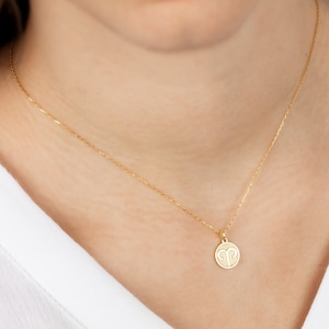 May include: A gold-colored necklace with a delicate chain and a round pendant. The pendant features the Aries zodiac symbol. The necklace is worn around the neck, against a white shirt.