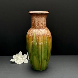 Puede incluir: Un jarrón de cerámica alto con una parte superior marrón moteada y un cuerpo verde texturizado. El jarrón tiene un borde acampanado y un cuello estrecho. Unas flores blancas están al lado del jarrón. El fondo es negro.