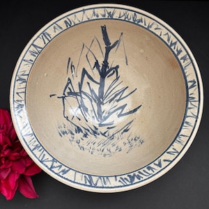 Puede incluir: Un cuenco de cerámica beige con un diseño de árbol abstracto azul en el centro. El borde presenta un patrón geométrico azul. El cuenco es probablemente hecho a mano y adecuado para servir alimentos o como pieza decorativa.