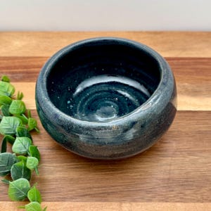 Könnte beinhalten: Eine handgefertigte, dunkelblaue Keramikschale mit gesprenkeltem Innenraum. Die Schale hat eine abgerundete Form und eine glänzende Oberfläche. Die Außenseite hat ein leicht strukturiertes Aussehen. Die Schale wird auf einer Holzoberfläche präsentiert.