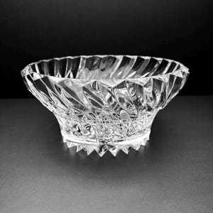 May include: A clear, faceted glass bowl with a wide, open top and a textured, geometric design. The bowl has a low profile and sits on a flat base with pointed edges. The bowl is designed to catch and reflect light.